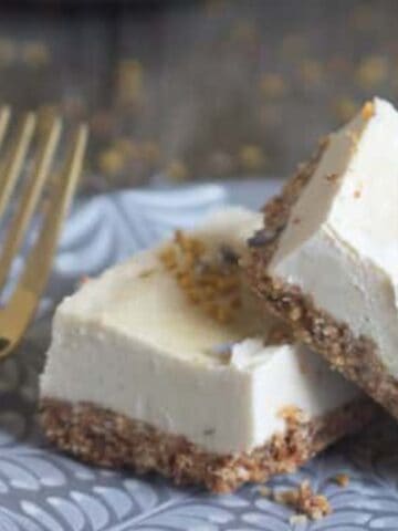 Two cheesecake bars with a crumb crust are placed on a decorative gray plate. The bars are topped with a sprinkle of crumble. A gold fork rests beside them on the plate. The background is a wooden surface with scattered crumbs.