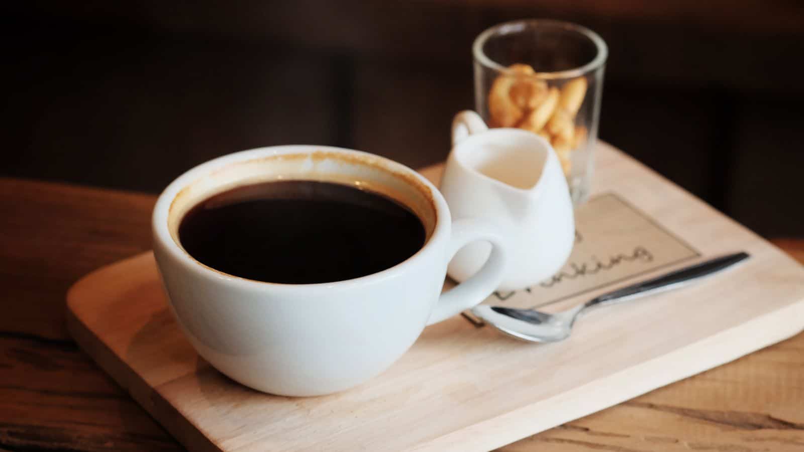 A cup of black coffee is placed on a wooden tray alongside a small white creamer pitcher and a glass containing a few nuts. A silver spoon rests on the tray, and a small piece of paper with writing is partially visible.