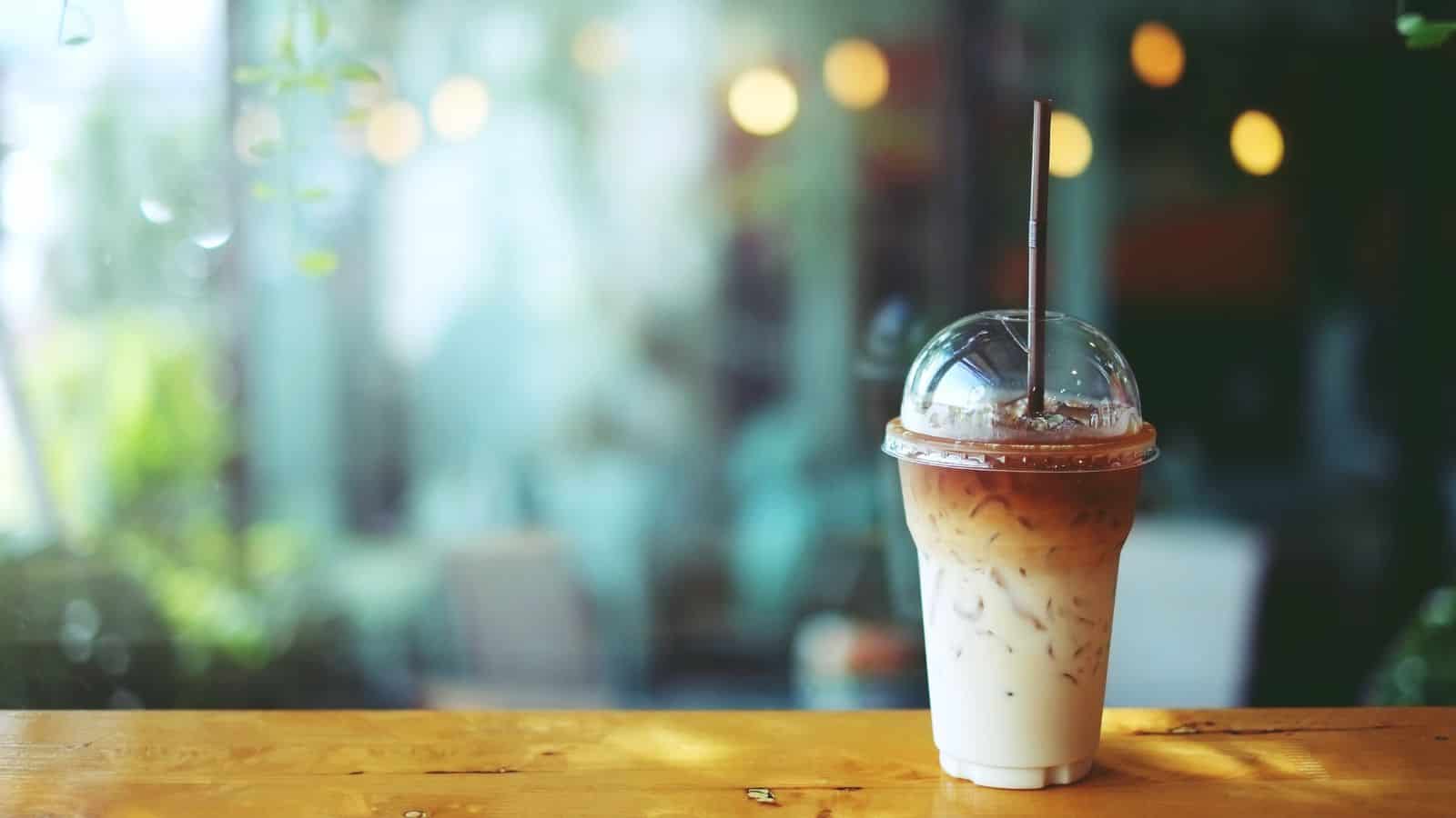A plastic cup of iced coffee with a straw sits on a wooden table. The drink has a gradient color, with darker coffee on top and milk below. In the background, there are blurred lights and greenery, suggesting a cozy cafe setting.