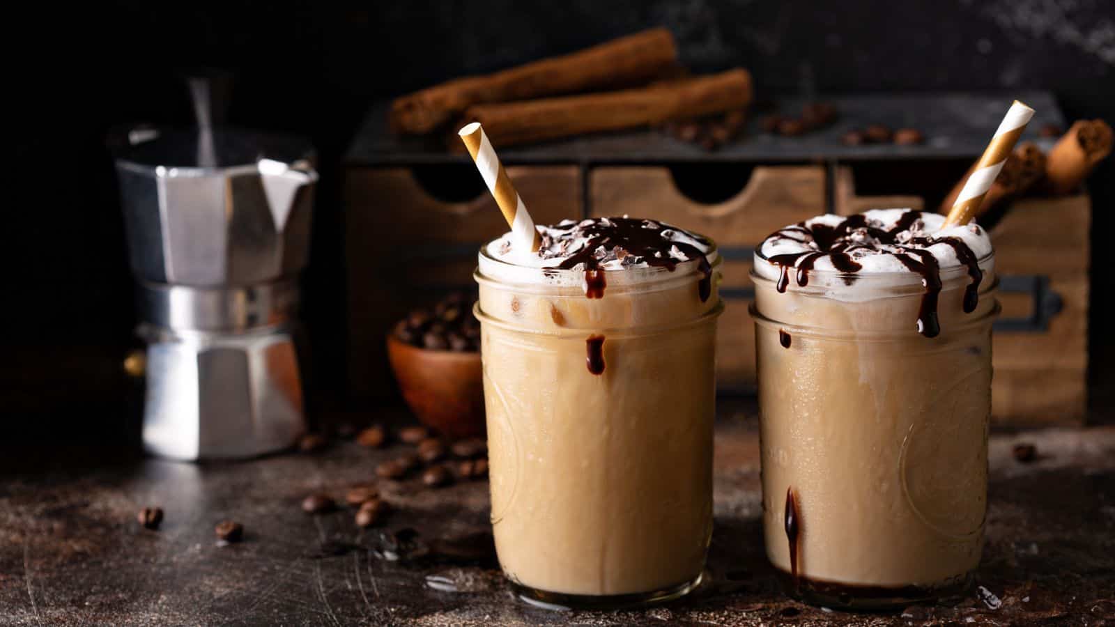 Two glass jars filled with iced coffee topped with whipped cream and chocolate drizzle, accompanied by striped straws. In the background are a moka pot, coffee beans, and a wooden drawer set, with cinnamon sticks scattered around.
