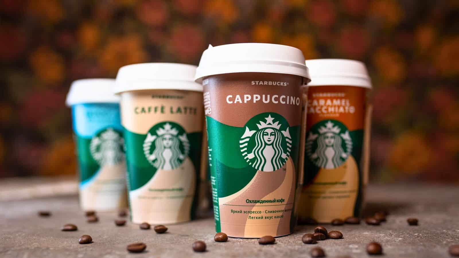 Four Starbucks coffee cups are arranged in a row with coffee beans scattered around them. The visible flavors include Cappuccino, Caff&egrave; Latte, and Caramel Macchiato. Each cup features the Starbucks logo and a distinctive color scheme.