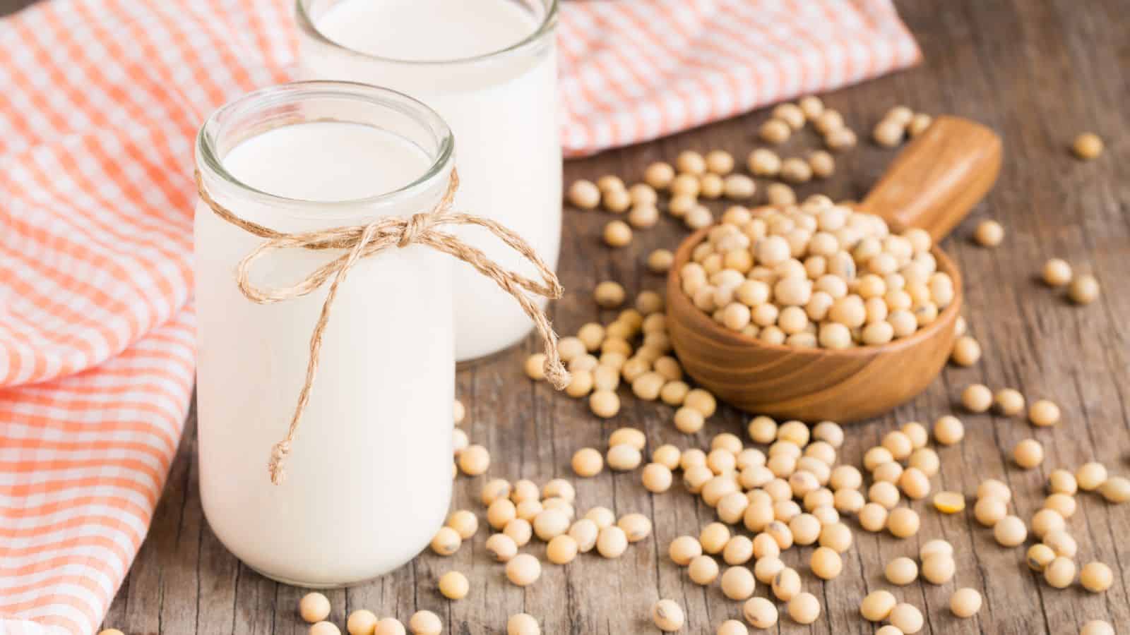 Two glass jars filled with soy milk, one tied with twine, are placed on a wooden surface. To the right, a small wooden scoop holds soybeans with more scattered around. An orange and white checkered cloth is partially visible in the background.