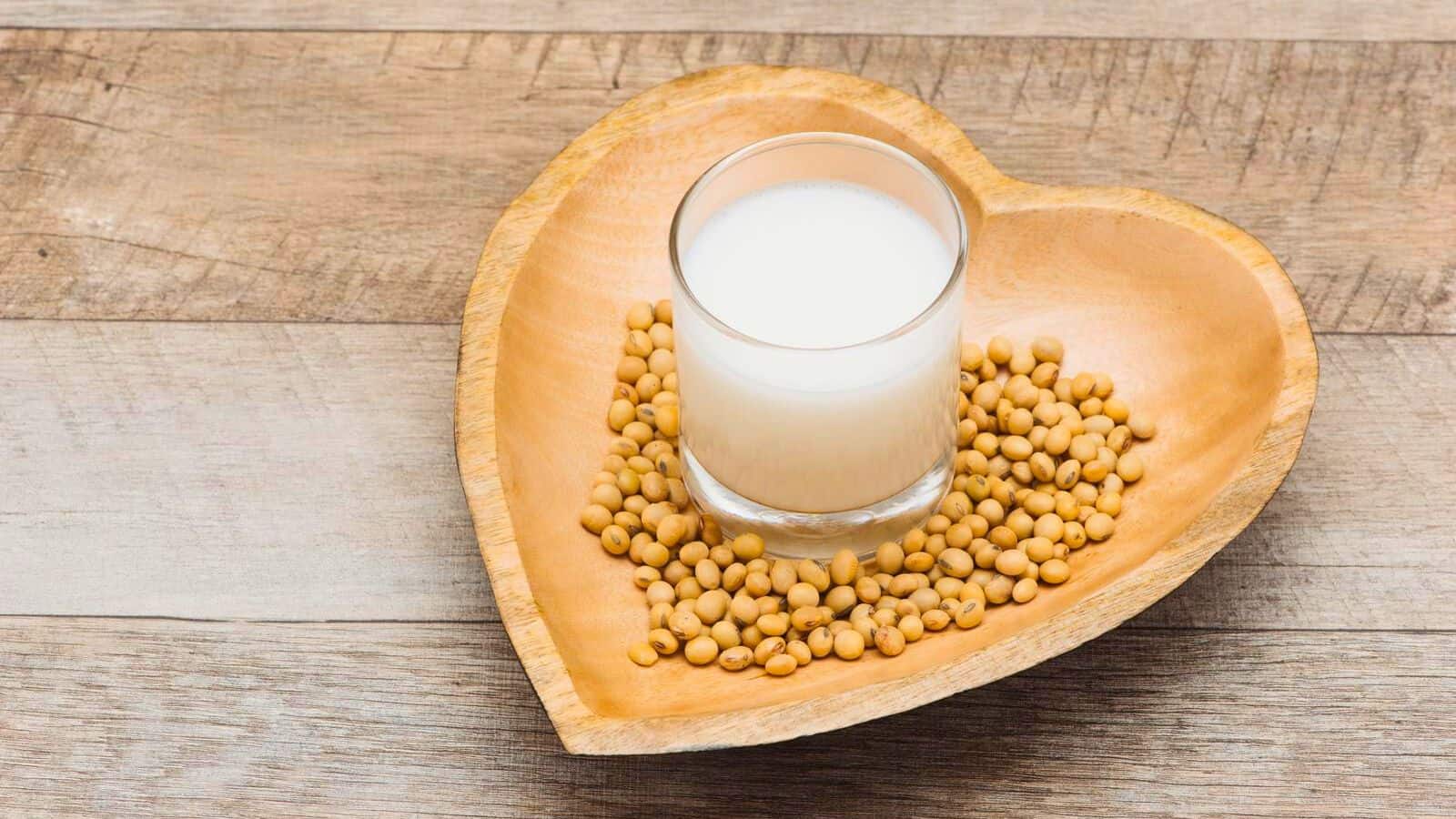 A glass of soy milk sits on a heart-shaped wooden tray surrounded by soybeans. The tray is placed on a wooden surface.