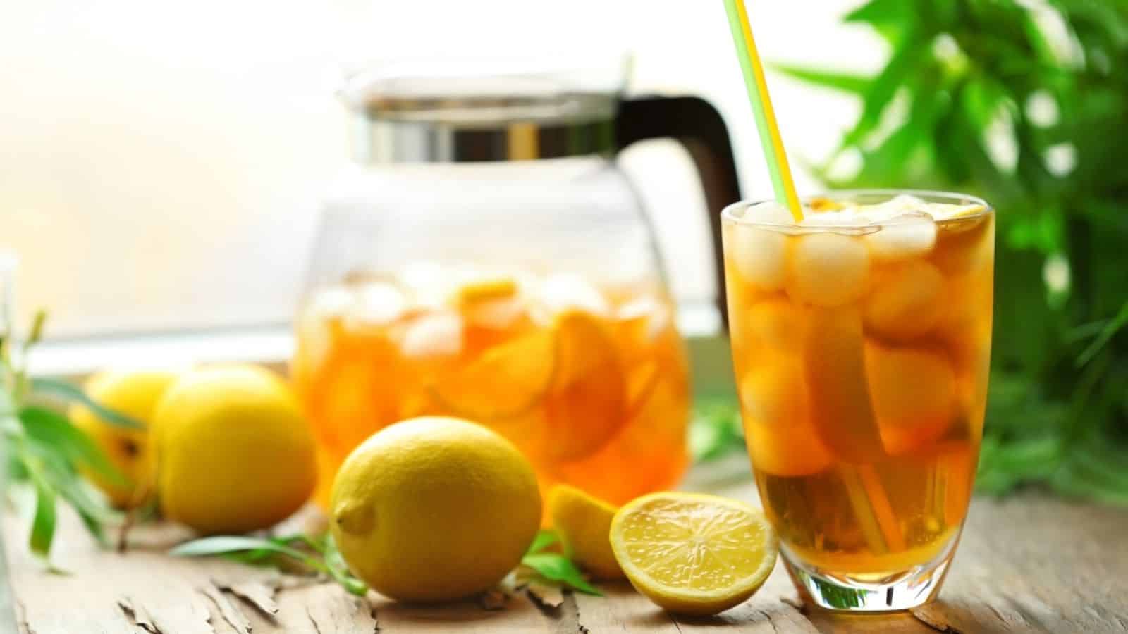 A glass of iced tea with lemon slices and a yellow straw is in the foreground, surrounded by whole and halved lemons. In the background, there is a glass pitcher of iced tea. Greenery is also visible, adding a fresh look to the scene.