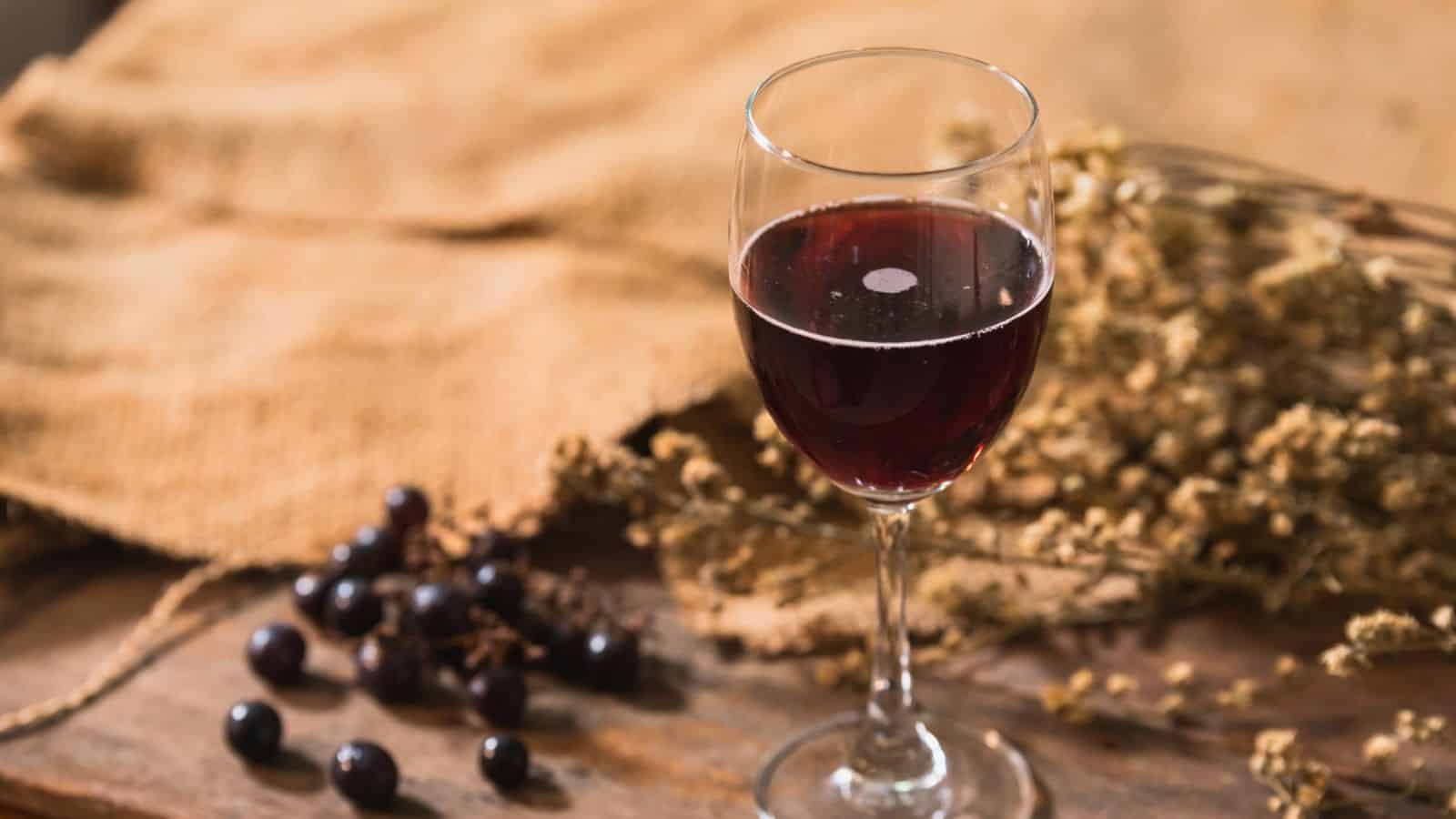 A glass of red wine sits on a wooden table alongside a cluster of dark grapes. In the background, there is a piece of burlap fabric and dried flowers, creating a rustic setting.