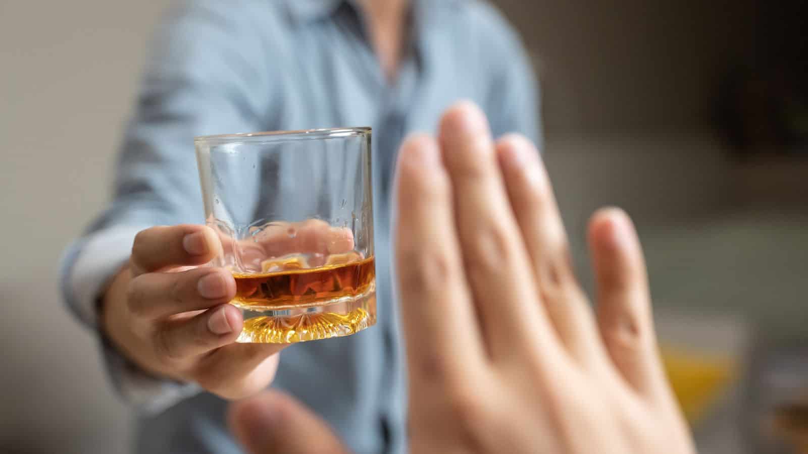 A person in a blue shirt is offering a glass of amber liquid to another person who is gesturing with their hand to refuse it. The focus is on the glass and the rejecting hand in the foreground.
