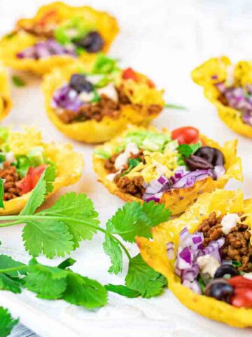 Mini taco cups made with crispy shells filled with seasoned ground meat, shredded lettuce, chopped tomatoes, black olives, diced onions, and cheese are arranged on a white platter. Fresh cilantro is placed alongside as garnish.