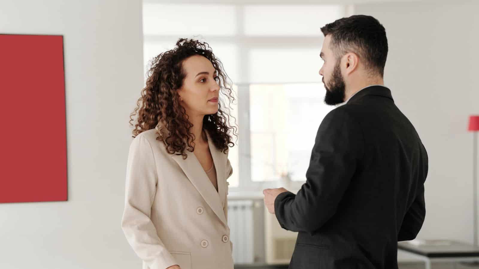 A woman with curly hair in a beige suit listens attentively to a man with a beard in a black suit. They are standing in a bright room with a large window, a red square artwork on the wall, and a pink floor lamp in the background.