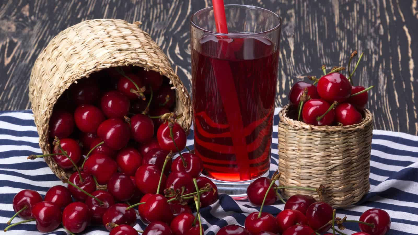 A glass of cherry juice with a red straw is placed on a black and white striped cloth. Next to the glass are two woven baskets, one tipped over, spilling cherries, and the other filled with cherries. The background is a dark wooden surface.
