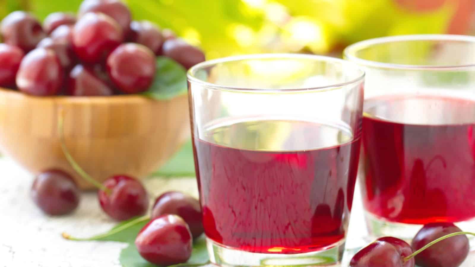 Two glasses of cherry juice are on a white surface, accompanied by fresh cherries scattered around. A wooden bowl filled with cherries is in the background, partially blurred. Bright green leaves are faintly visible.
