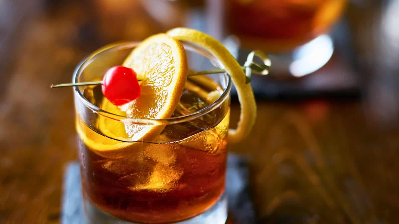 A glass filled with a dark cocktail sits on a wooden table. The drink is garnished with an orange slice, a maraschino cherry, and a lemon twist. Ice cubes are visible in the glass. A blurred background shows another similar drink.