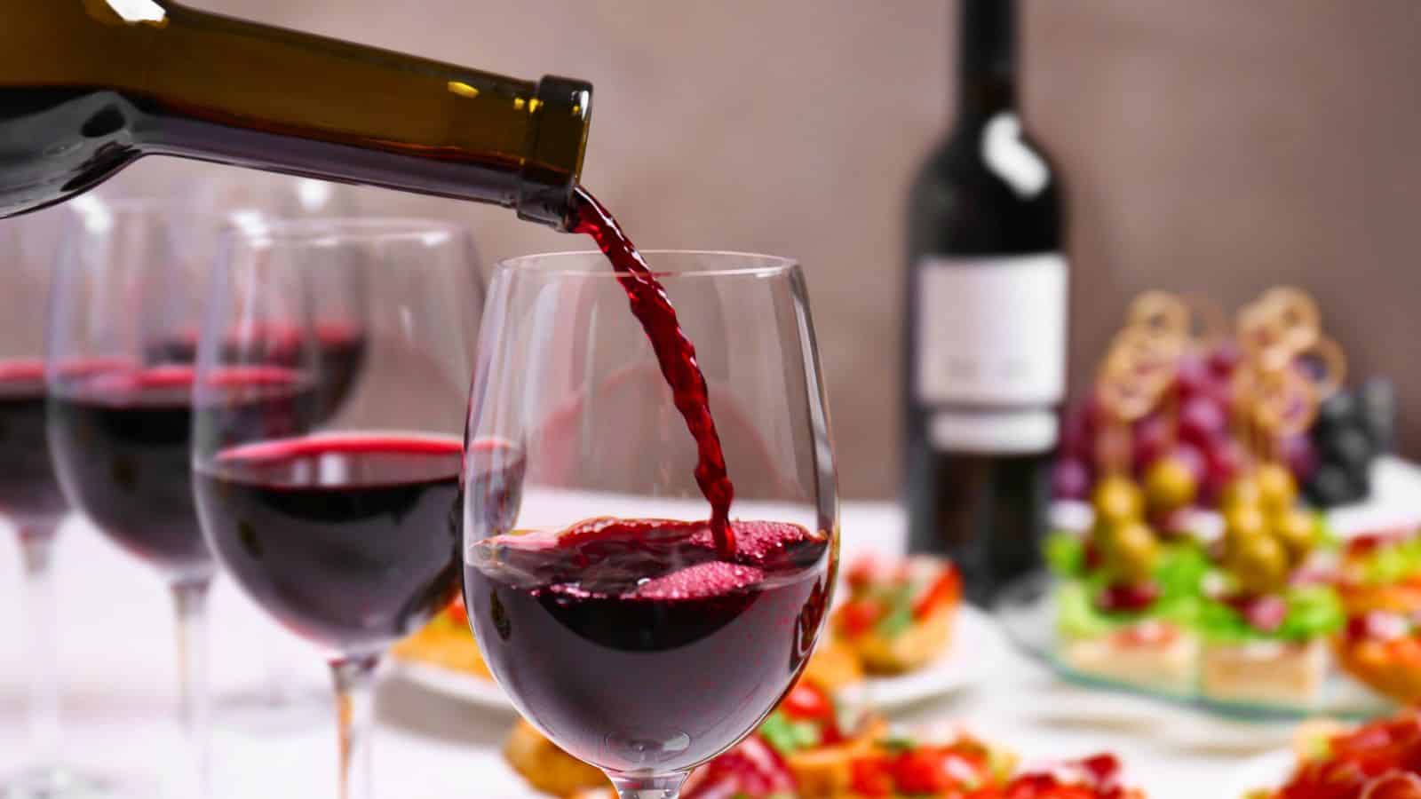 A bottle pours red wine into a glass set on a table. Other filled wine glasses and a variety of food, including grapes and appetizers, are in the background. A blurred wine bottle is also visible.