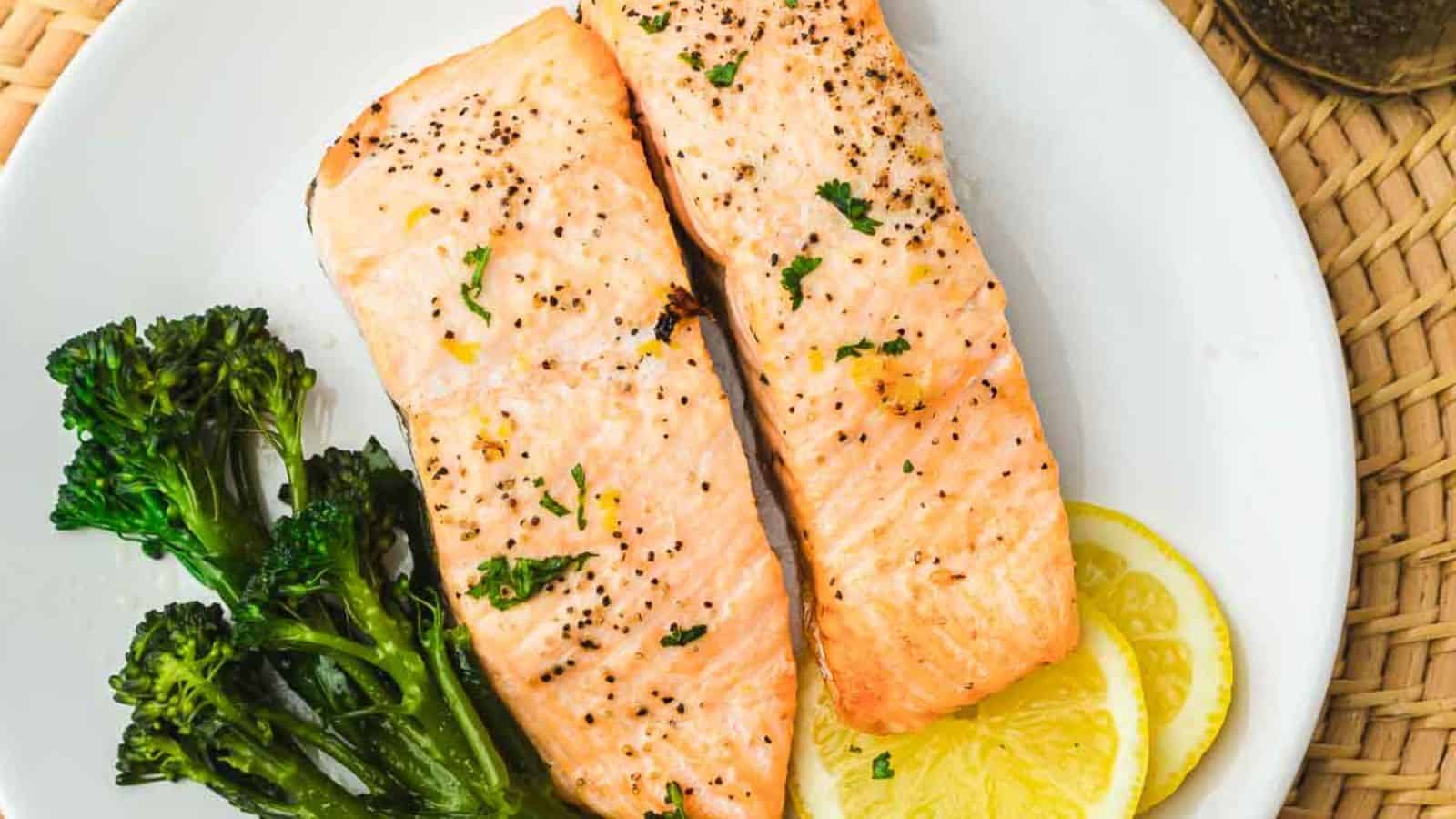 Four grilled salmon fillets garnished with fresh dill and lemon slices are arranged on a white oval platter. The fillets are seasoned with herbs and pepper. The edge of a striped cloth and a serving of broccoli are partially visible.