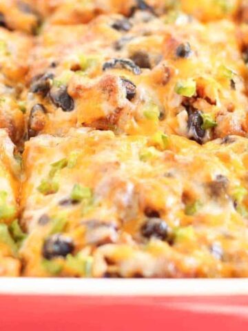 Close-up of a baked casserole in a dish, topped with melted cheese, black beans, and chopped green onions. The dish appears hot and freshly cooked, with golden brown cheese and visible ingredients.