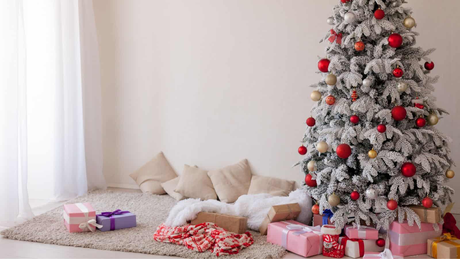 A Christmas tree decorated with red and gold ornaments stands in a corner of a room. Several wrapped gifts in various sizes are placed beneath the tree. A few beige cushions and a blanket are on the floor nearby.