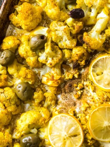 A baked dish featuring roasted cauliflower florets seasoned with spices, garnished with slices of lemon and green olives. A teal bowl with green olives and a small bowl with a dark sauce are seen in the background.