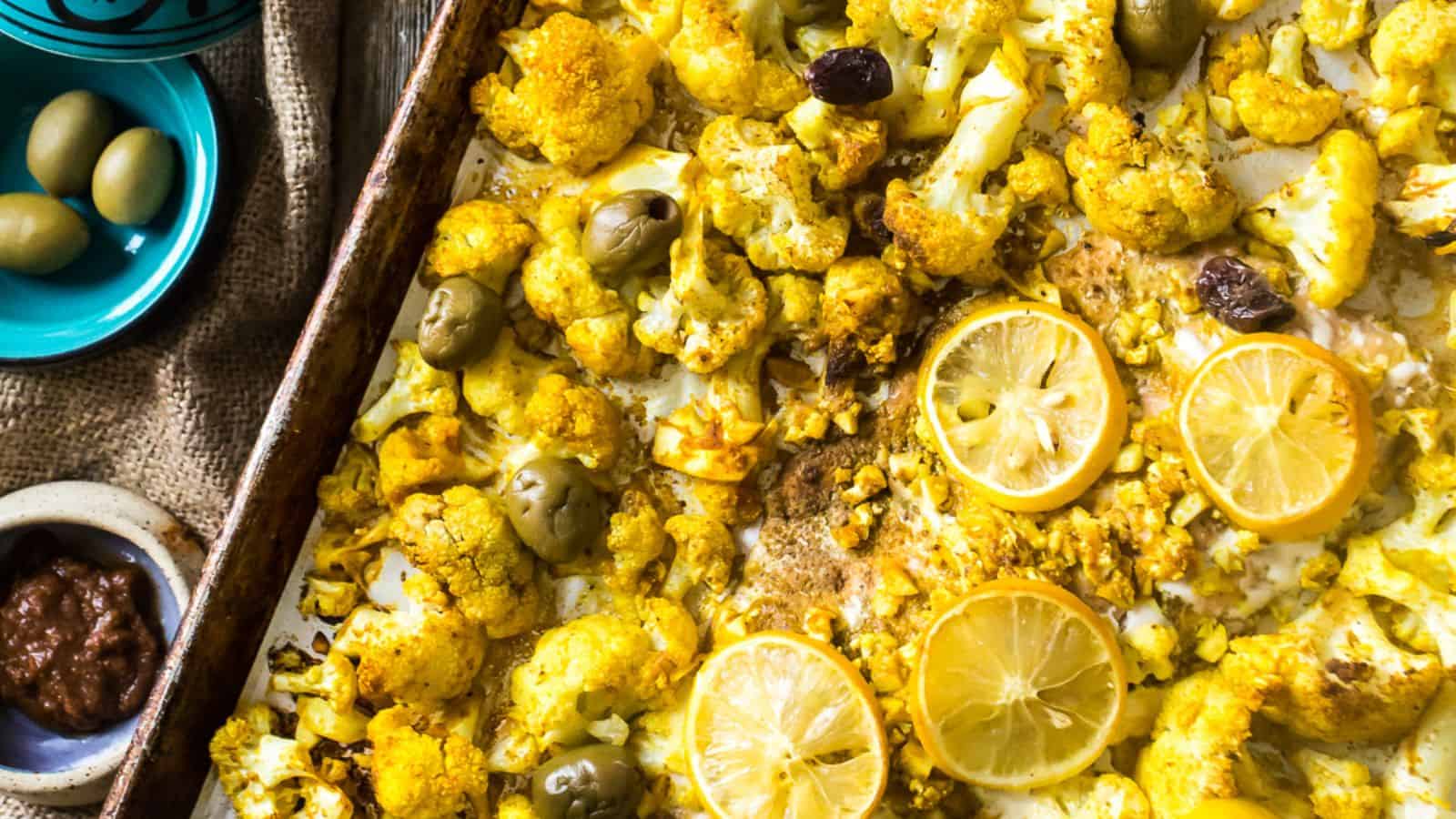 A baking tray displays roasted cauliflower florets, lemon slices, and olives alongside a piece of salmon. A small bowl of olives and another with a dark sauce are placed nearby on a burlap-covered surface.