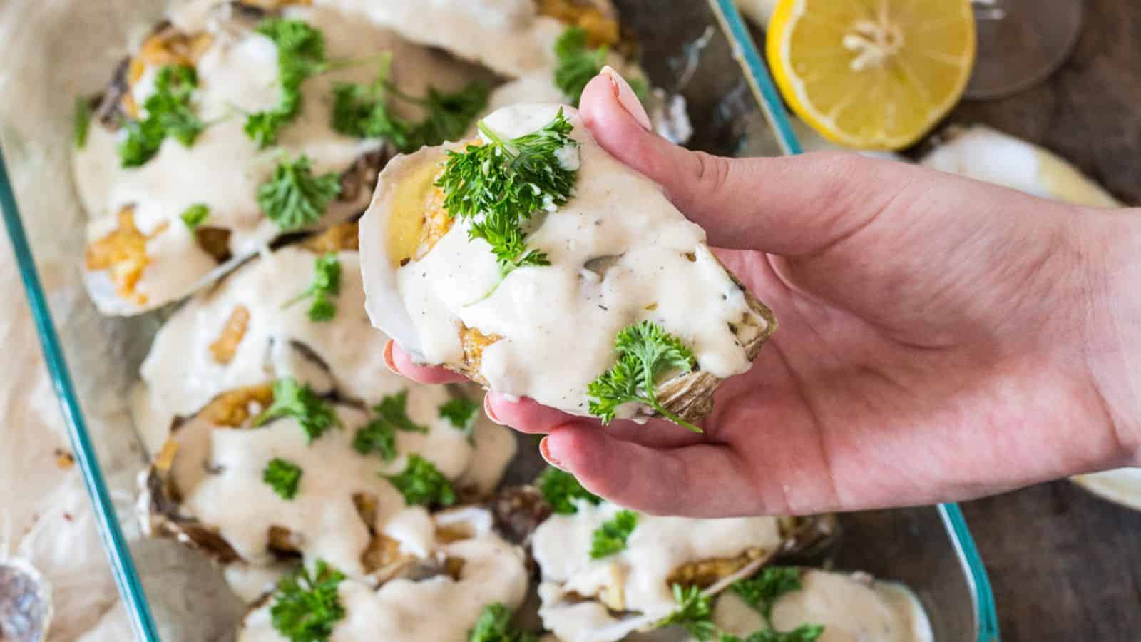 A hand holding a baked oyster topped with creamy sauce and garnished with parsley. Several similar oysters are placed in a glass tray below. A slice of lemon and a glass are visible in the background.