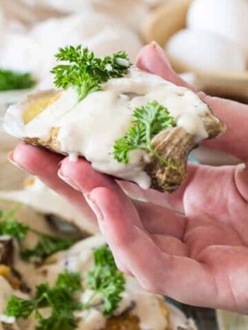 A hand holding a potato slice topped with creamy white sauce and garnished with fresh parsley. In the background, there are more similar potato slices, a bowl of eggs, and some parsley.