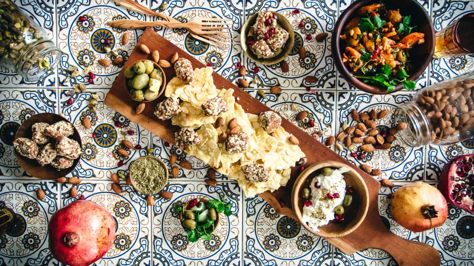 An overhead view of a colorful spread on a tiled table. It includes a wooden board with cheese and crackers, bowls of olives, salad, nuts, and dips. A pomegranate and almonds are also scattered around for decoration.
