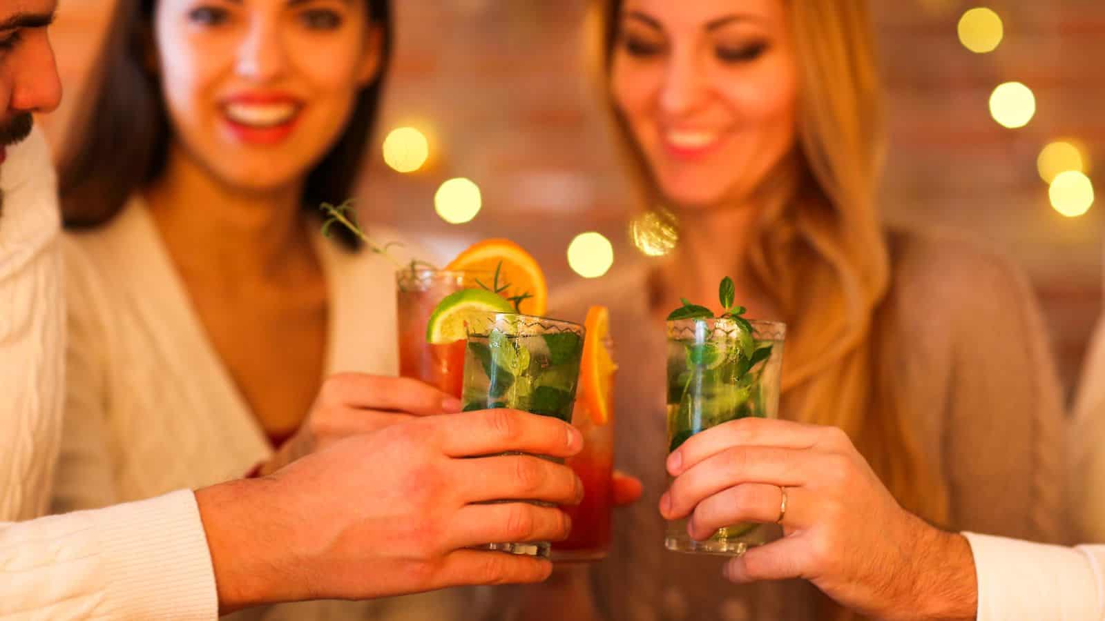 A group of people clinks glasses filled with colorful drinks, garnished with herbs and fruit slices. The background is softly lit, with bokeh lights creating a festive atmosphere. The scene suggests a social or celebratory gathering.