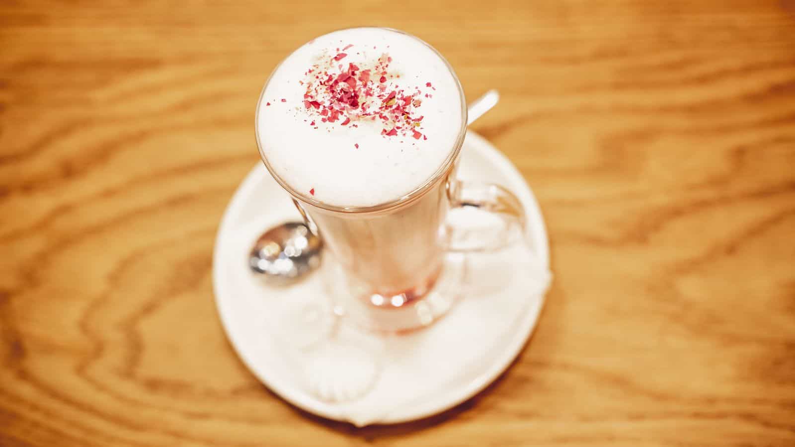A tall glass of frothy latte topped with pink sprinkles is placed on a white saucer with a teaspoon beside it, all set on a wooden table.