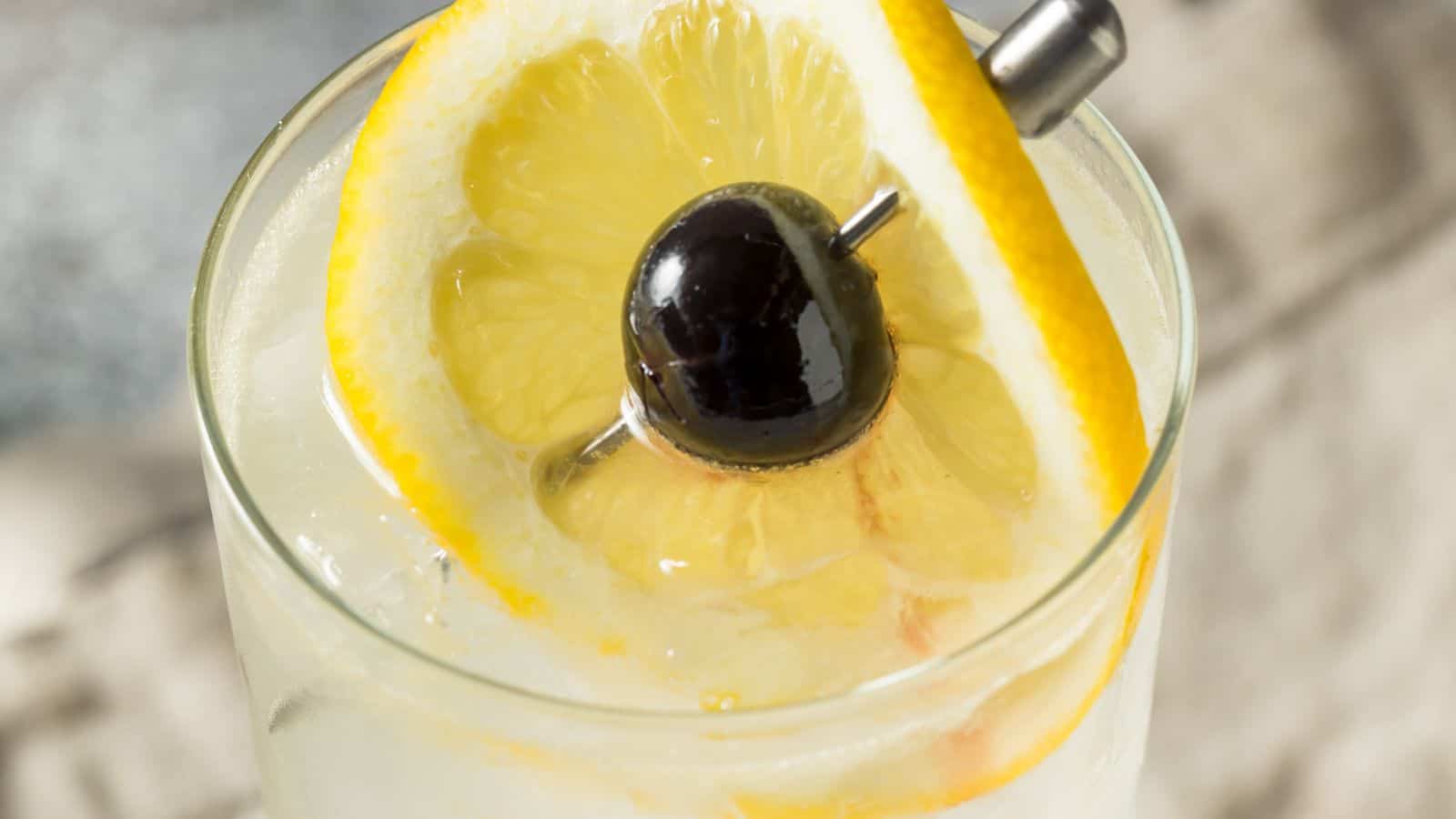 A close-up of a cocktail in a glass garnished with a slice of lemon and a dark cherry on a metal pick. The drink has a light, translucent appearance and is set against a blurred background.