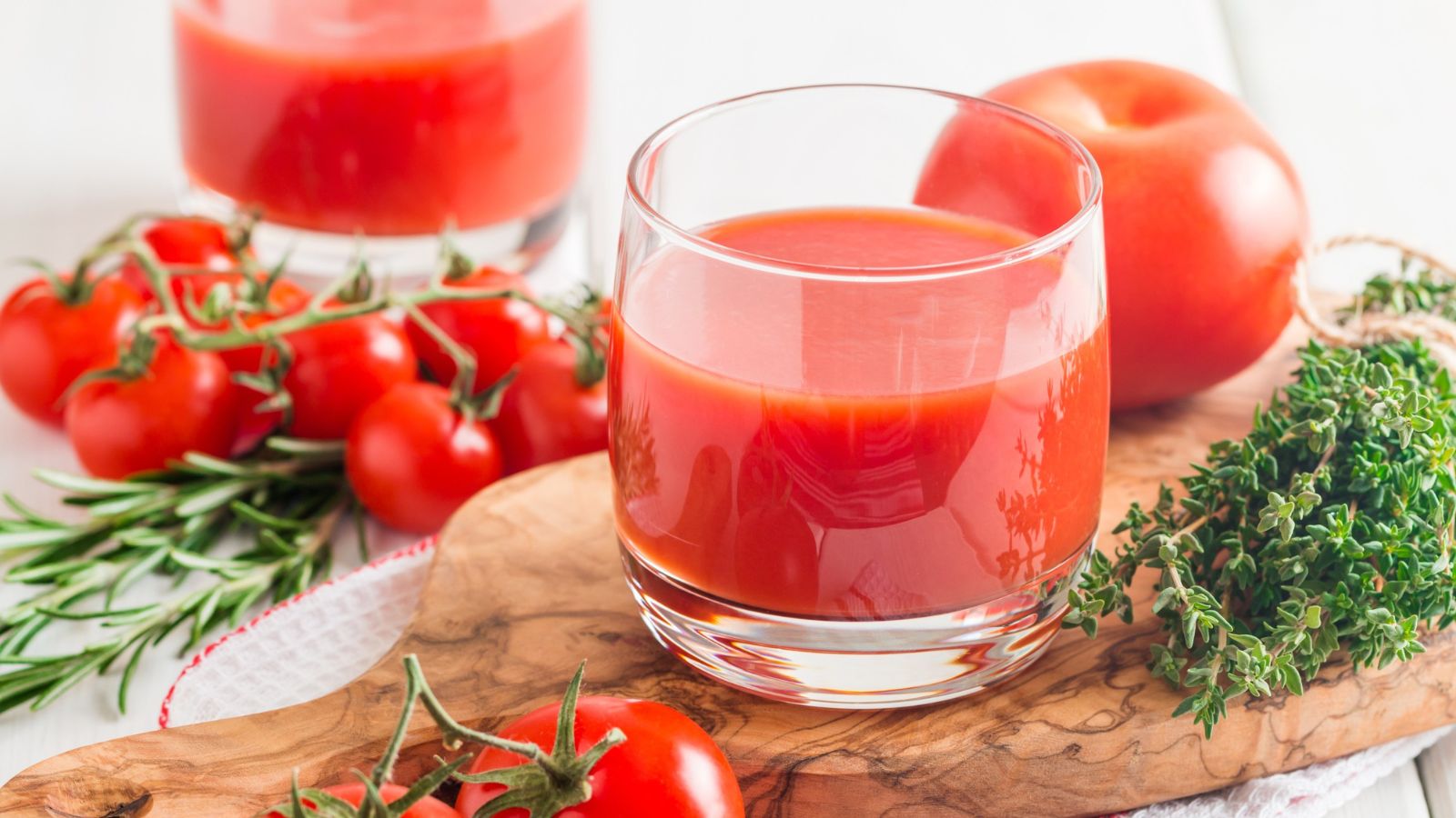 A glass of tomato juice is placed on a wooden board, surrounded by fresh tomatoes, rosemary, and thyme. Another glass of juice and more tomatoes are in the background on a white surface.