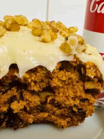 A slice of cake with cream cheese frosting topped with walnuts on a plate. A fork is beside it. In the background, there is a can of Campbell's tomato soup.