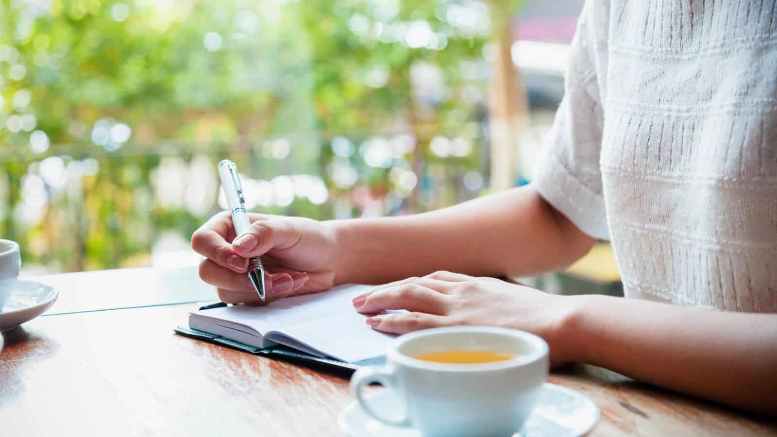 A person sits at a wooden table, writing in a notebook with a pen. A cup of tea is placed nearby. The background features blurred greenery, suggesting an outdoor setting or view through a window.