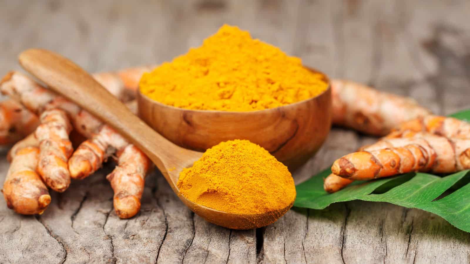 A wooden bowl and spoon filled with bright yellow turmeric powder are placed on a rustic wooden surface. Fresh turmeric roots are scattered around, along with a green leaf. The scene highlights natural and earthy textures and colors.