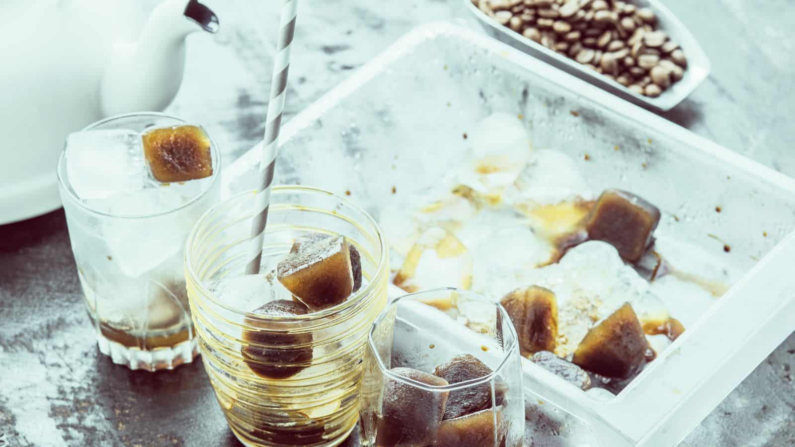 Glasses filled with coffee ice cubes sit on a tabletop next to a tray of more cubes. A teapot is partially visible on the left, and a bowl of coffee beans is in the background. A striped straw is in one of the glasses.