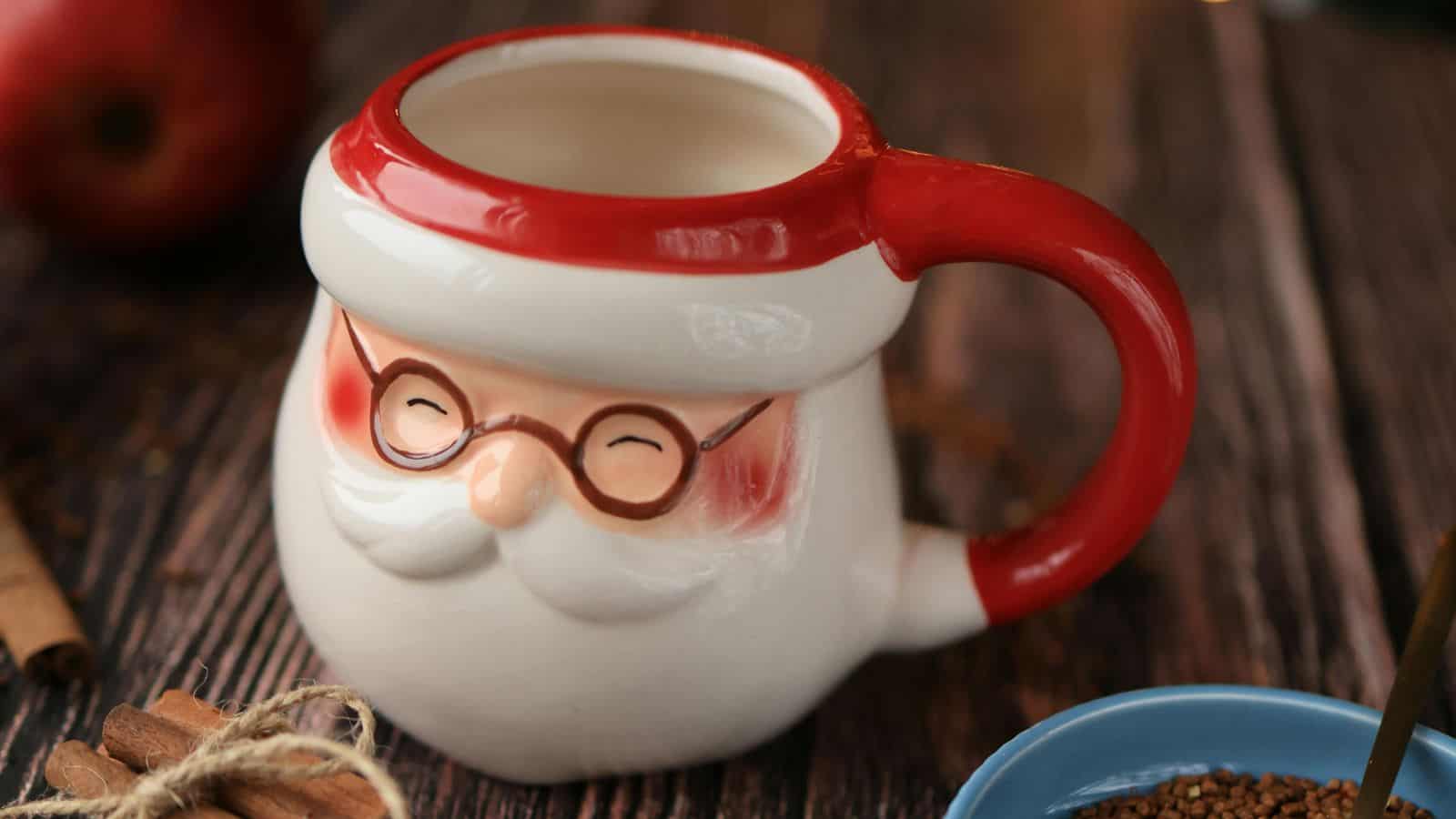 A ceramic mug shaped like Santa Claus with a red handle and a design featuring glasses, rosy cheeks, and a white beard is placed on a wooden table. Nearby are cinnamon sticks and an apple.