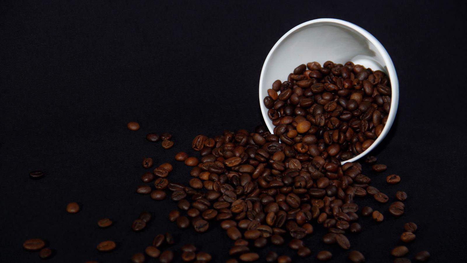 A white cup tipped over, spilling dark roasted coffee beans onto a black surface. The background is a solid black, highlighting the rich brown color of the beans.