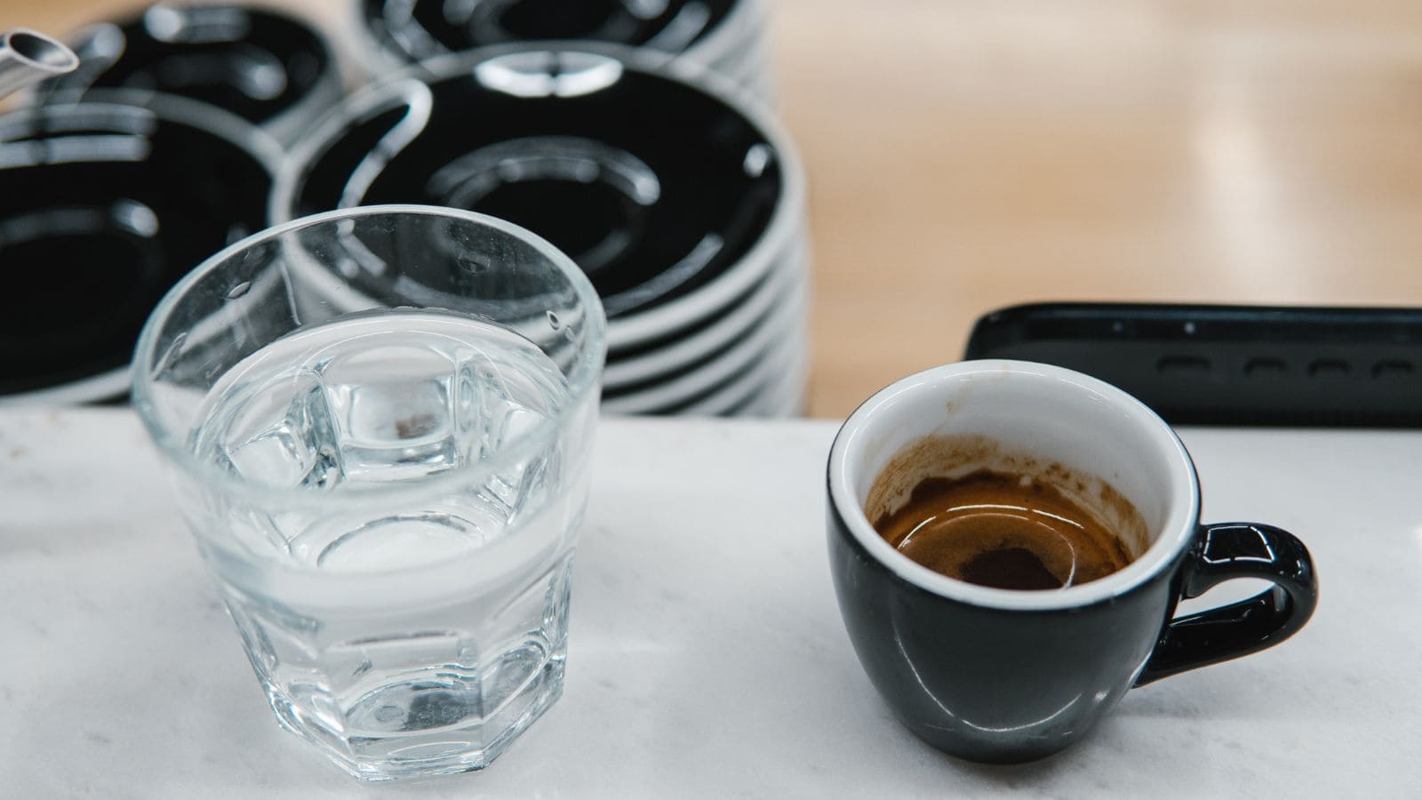 A small black espresso cup with coffee is on a countertop next to a clear glass of water. In the background, stacks of black and white saucers are visible.