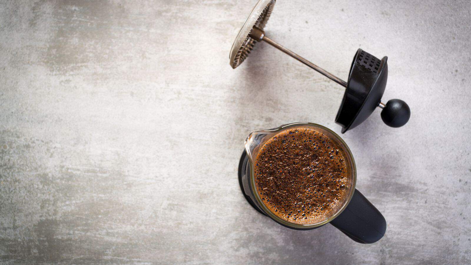 A French press filled with brewed coffee sits on a textured gray surface. The metal plunger is partially lifted and positioned beside the glass container. The coffee is dark and frothy on top.