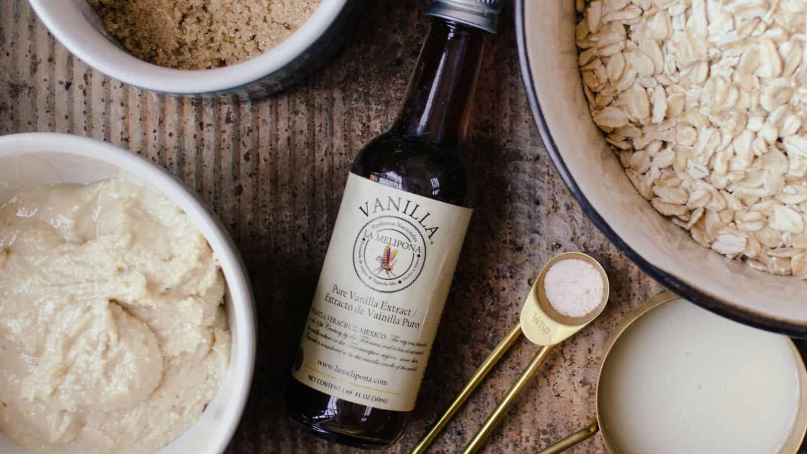 A bottle of vanilla extract lies on a wooden surface surrounded by bowls of brown sugar, oats, and a creamy mixture. A measuring spoon holds a light-colored powder next to a cup containing a small amount of milk.