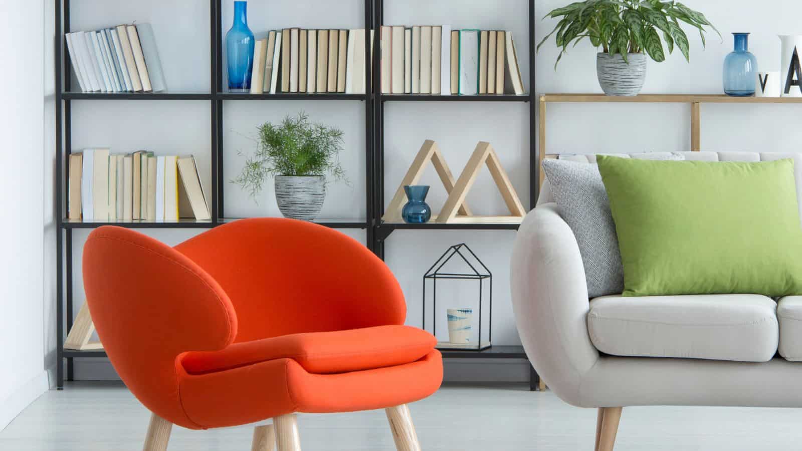 Modern living room with a bright orange chair and a beige sofa with gray and green cushions. A black bookshelf with books and decor items, including vases and plants, is in the background. The room has a minimalist and stylish design.