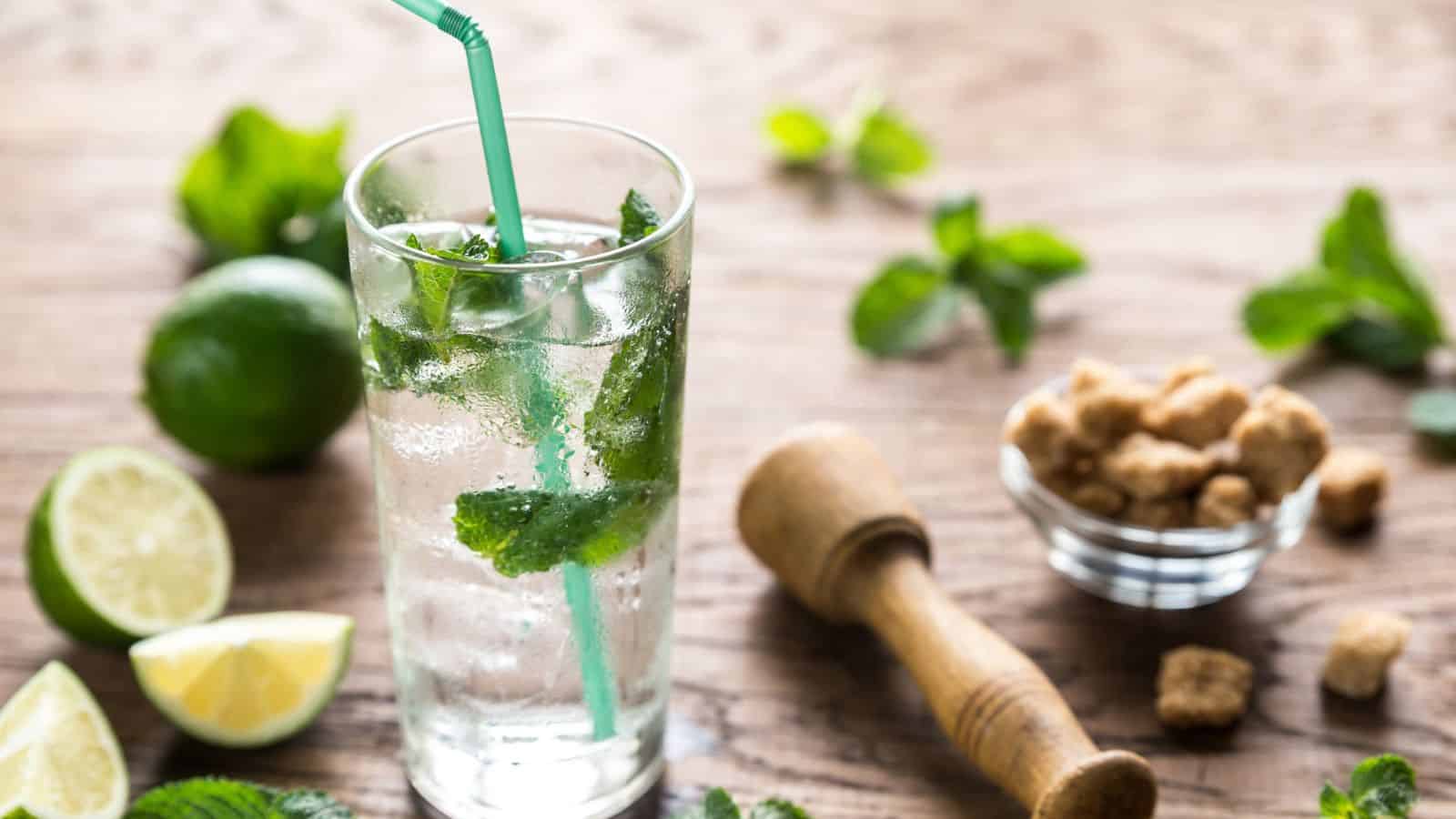 A glass of clear sparkling drink with mint leaves and a straw is on a wooden table. Surrounding the glass are lime wedges, a whole lime, fresh mint sprigs, a small bowl of brown sugar cubes, and a wooden muddler.