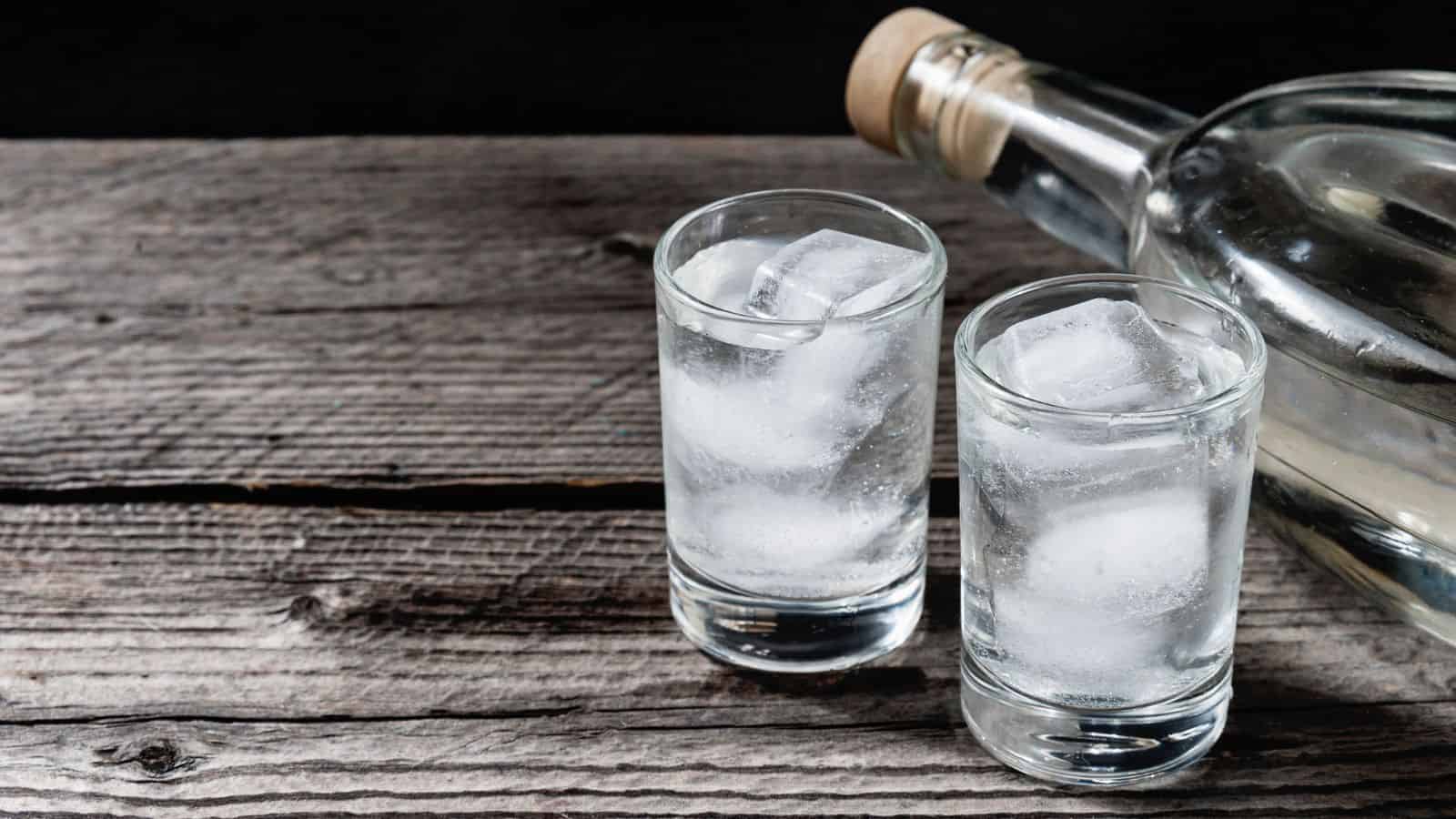 Two glasses filled with clear liquid and ice cubes are placed on a rustic wooden surface. A glass bottle with a cork stopper lies on its side nearby. The scene is set against a dark background.