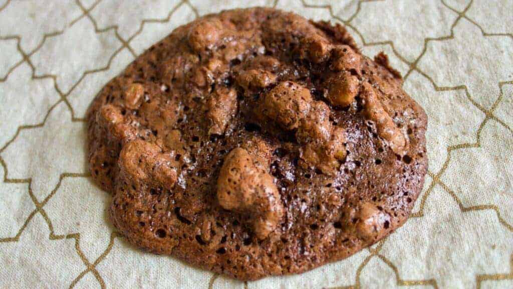 A round chocolate cookie with a rough, uneven texture and visible chunks, resting on a decorative background with a geometric pattern.