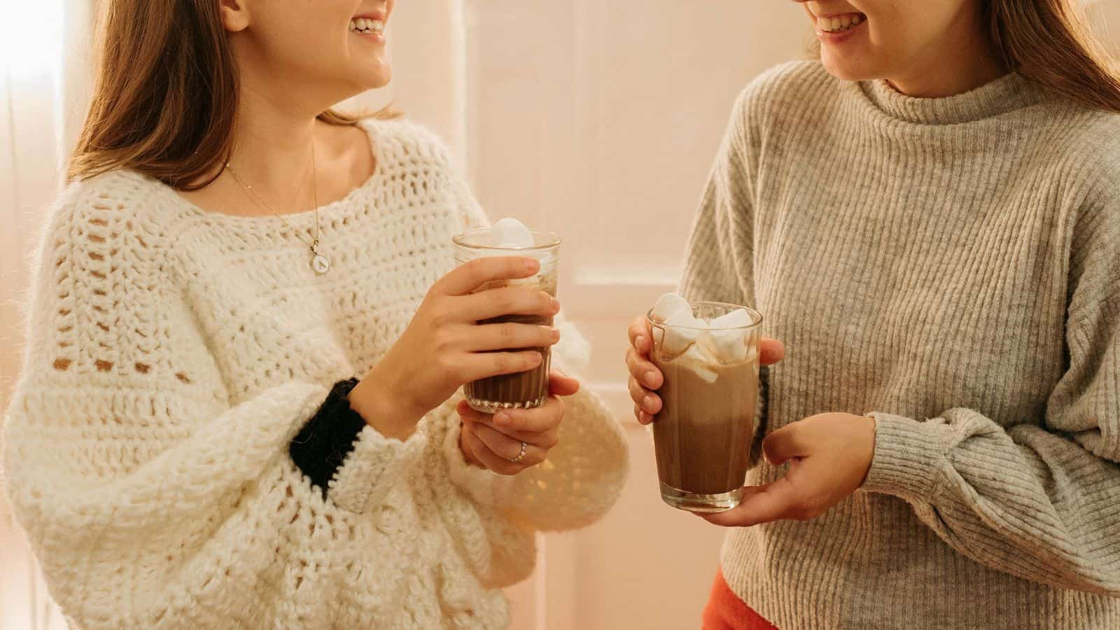 Two people are standing indoors, each holding a glass of hot chocolate topped with marshmallows. They are dressed in cozy sweaters, one white and one gray, and appear to be smiling and conversing.