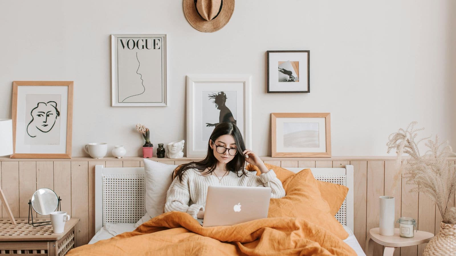 A person sits on a bed using a laptop, surrounded by yellow bed covers. The room features minimalist wall art, potted plants, a hat hanging on the wall, and a small round side table with a candle and books.