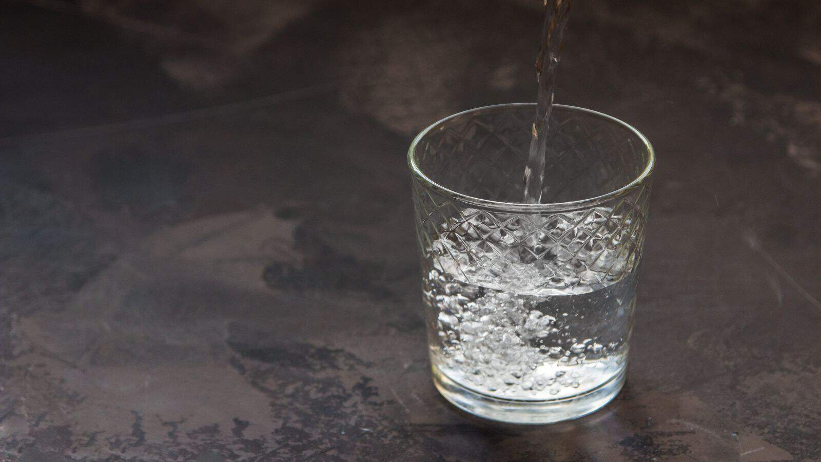 A clear glass on a dark surface is being filled with water. The glass has a textured, diamond-like pattern, and bubbles are visible as the water pours in.