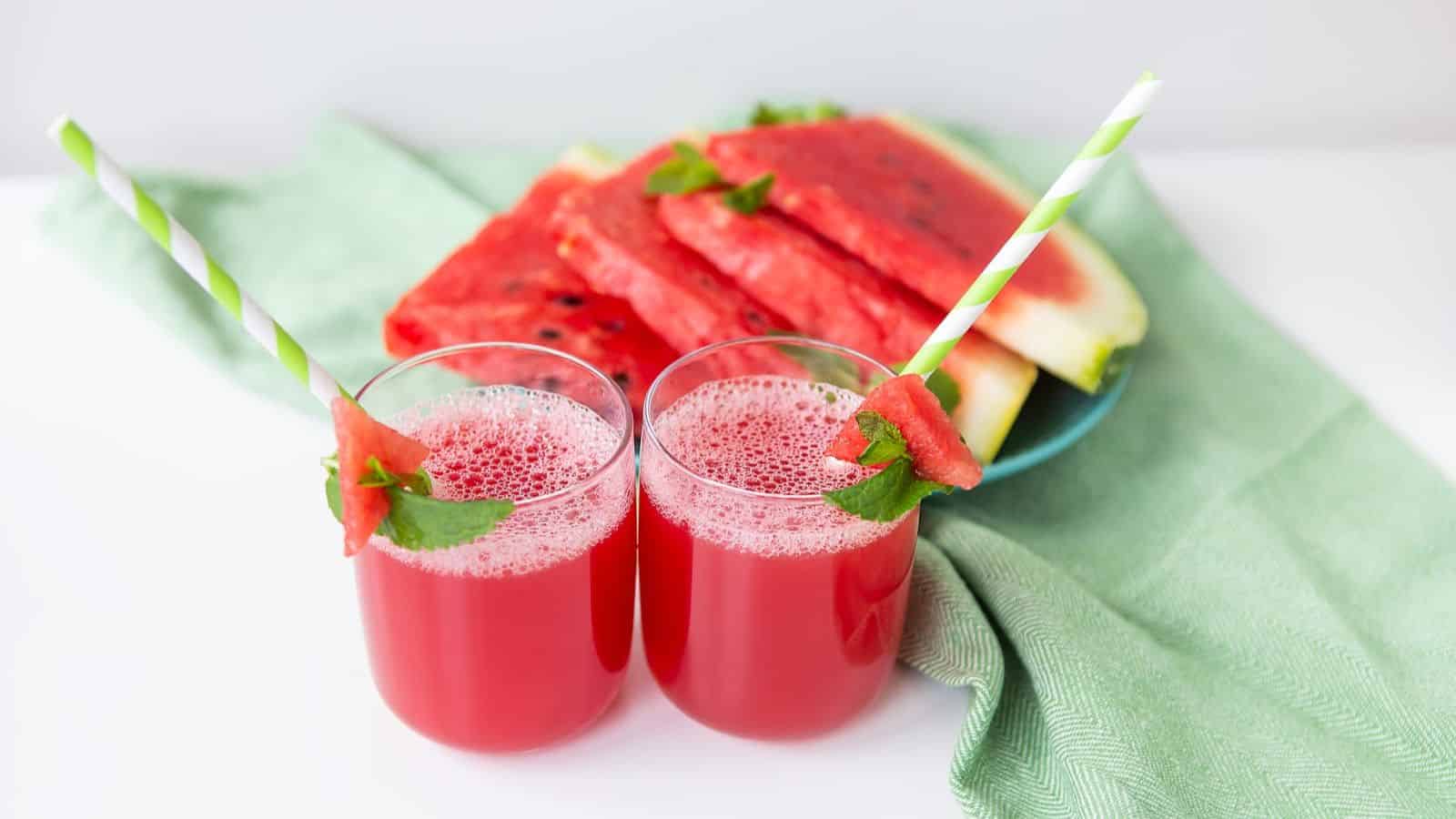 Two glasses of watermelon juice are garnished with mint and watermelon slices. Each glass has a green and white striped straw. In the background, there is a plate of watermelon slices on a green cloth.