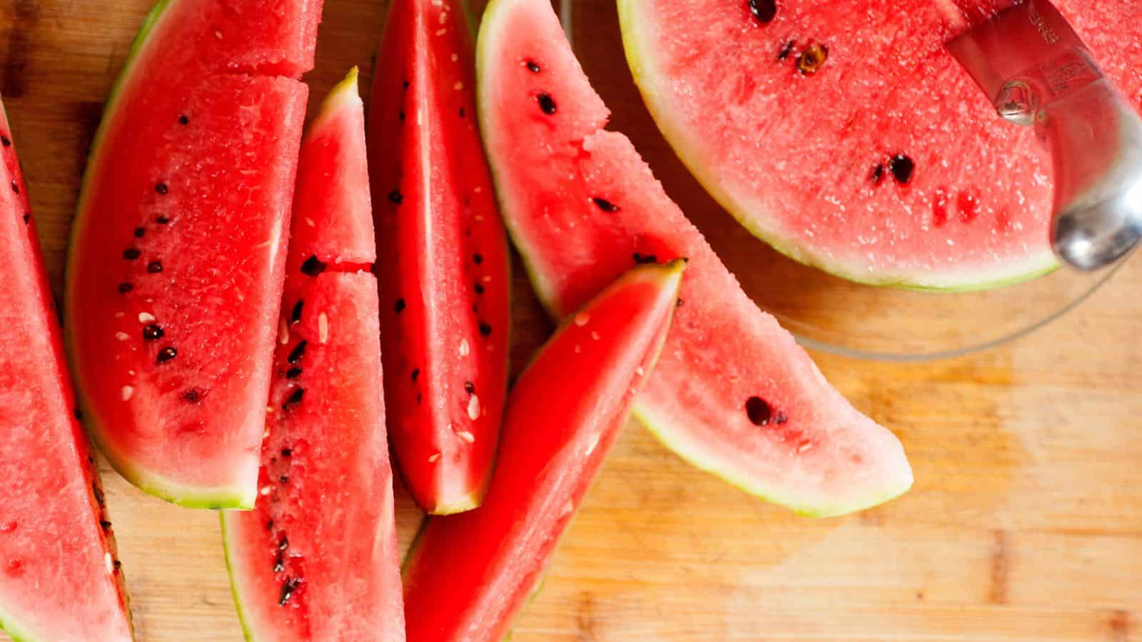 Sliced watermelon is arranged on a wooden cutting board. One larger piece is placed on a glass plate, and a metal kitchen tool is partially visible. The watermelon is bright red with dark seeds.