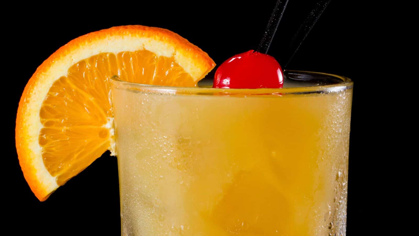 A close-up of a frosty cocktail in a glass, garnished with an orange slice and a red cherry on top, with two black straws inserted into the drink. The background is solid black.