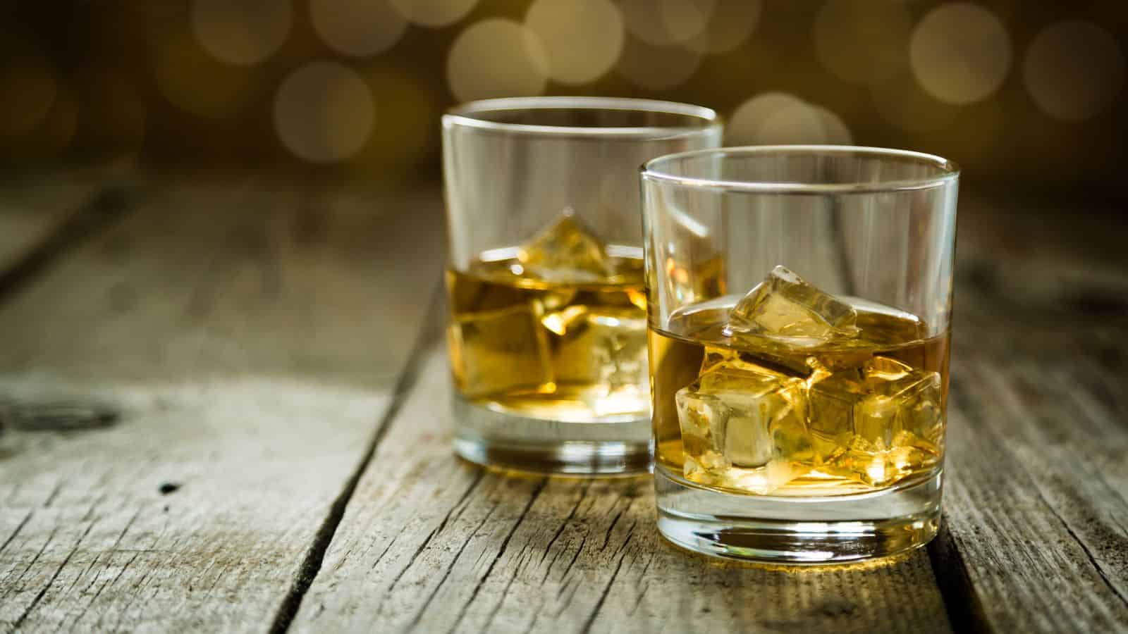 Two glasses filled with amber-colored liquid and ice cubes are placed on a rustic wooden table. The background features blurred, warm, circular bokeh lights, creating a cozy atmosphere.