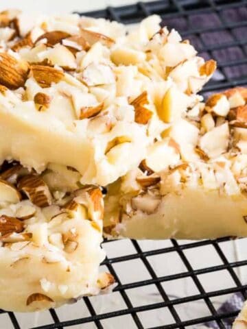 Squares of white chocolate fudge topped with chopped almonds are stacked on a black wire cooling rack. The fudge has a creamy appearance, and the almonds add texture and color. A dark cloth is partially visible beneath the rack.