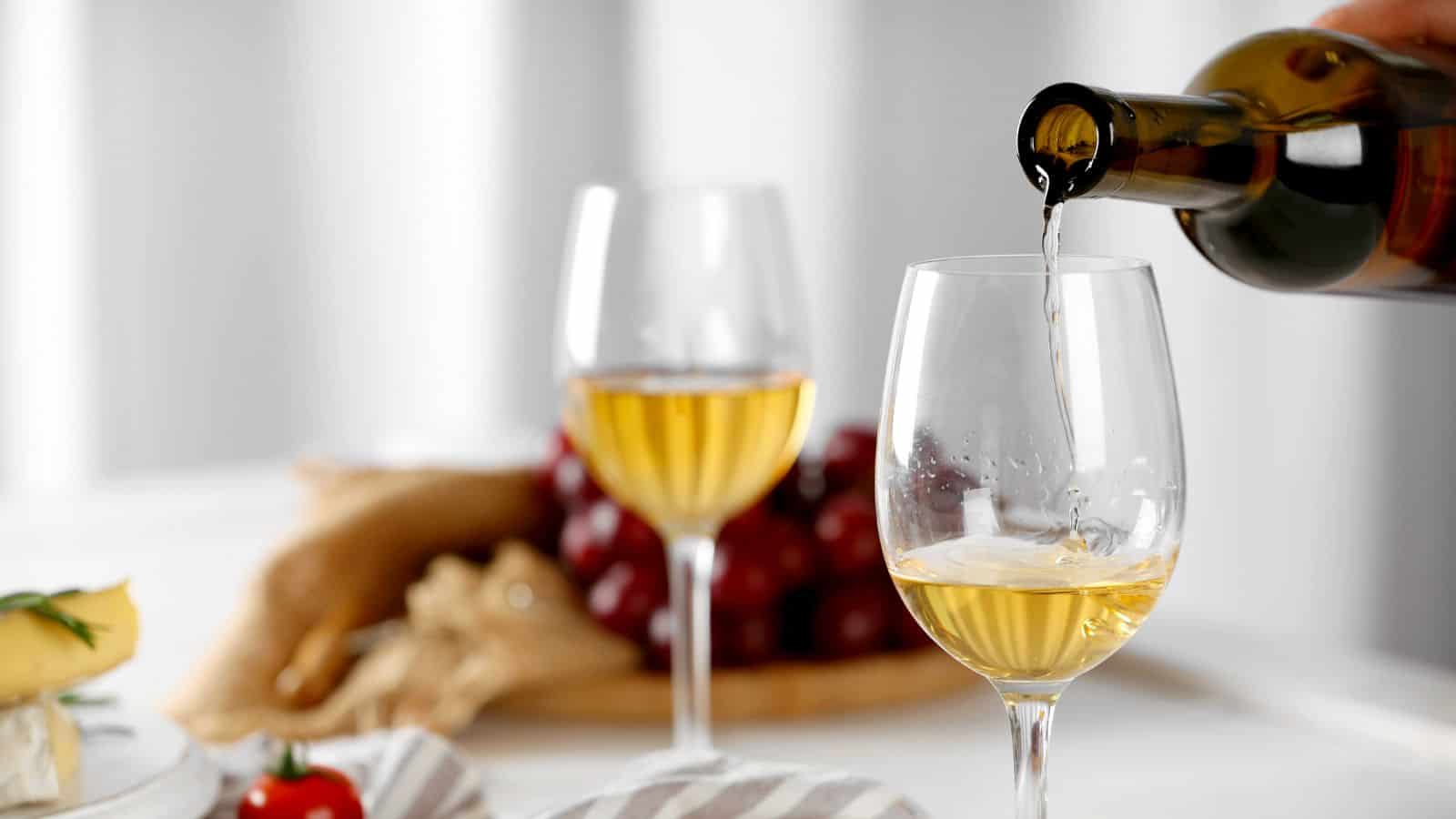 A close-up of white wine being poured into a glass. Another filled wine glass is in the background. A plate with grapes, cheese, and a tomato is partially visible on a table with a striped cloth.